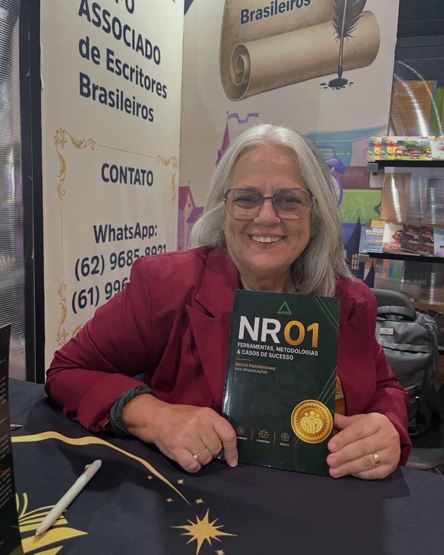 Dra. Cláudia Araújo sorrindo, segurando o livro NR01 no lançamento na Bienal do Livro de São Paulo.